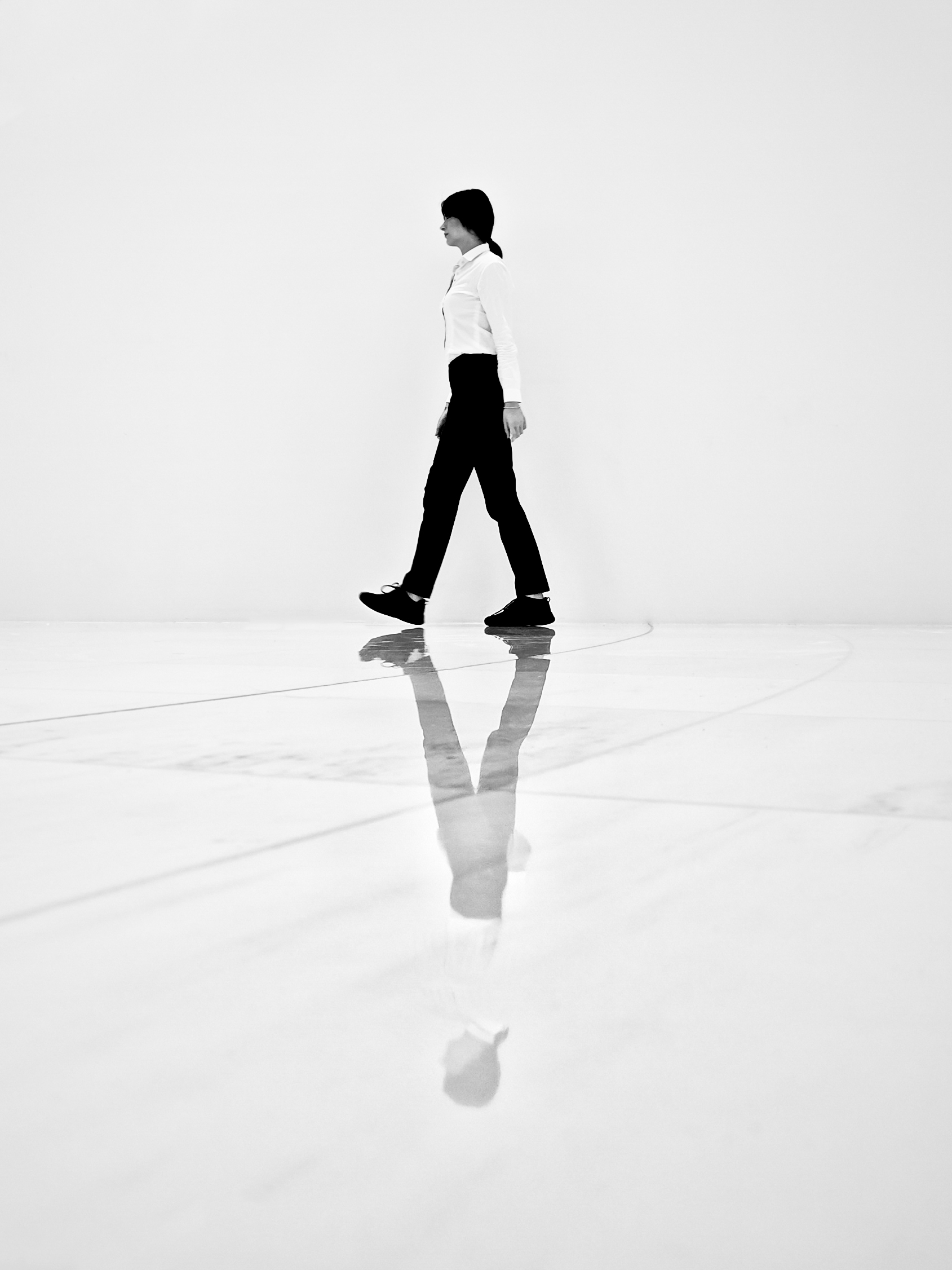 A woman in a white shirt and black trousers walks across a near-white polished plaza and is mirrored full-length in the wet marble; almost the entire frame is blank white