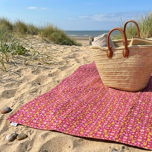 A woven straw French market basket with tan leather handles sits on a pink floral patterned picnic blanket spread on the sand of a dune-backed beach. Two pebbles are near the blanket's edge.