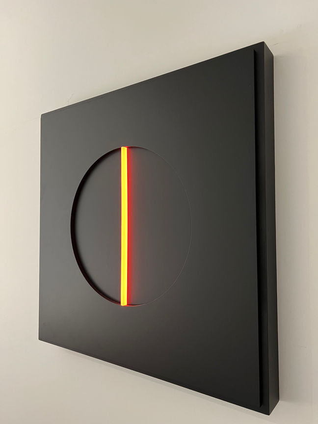 Black square light-art with an orange illuminated acrylic rod in deepened black circle.