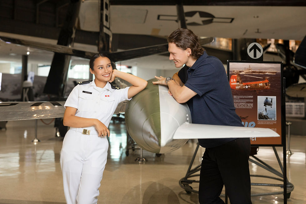 Two people in a museum lean on a plane nose, smiling at each other. One in a white naval uniform, the other in a blue polo. Aircrafts and displays in background.