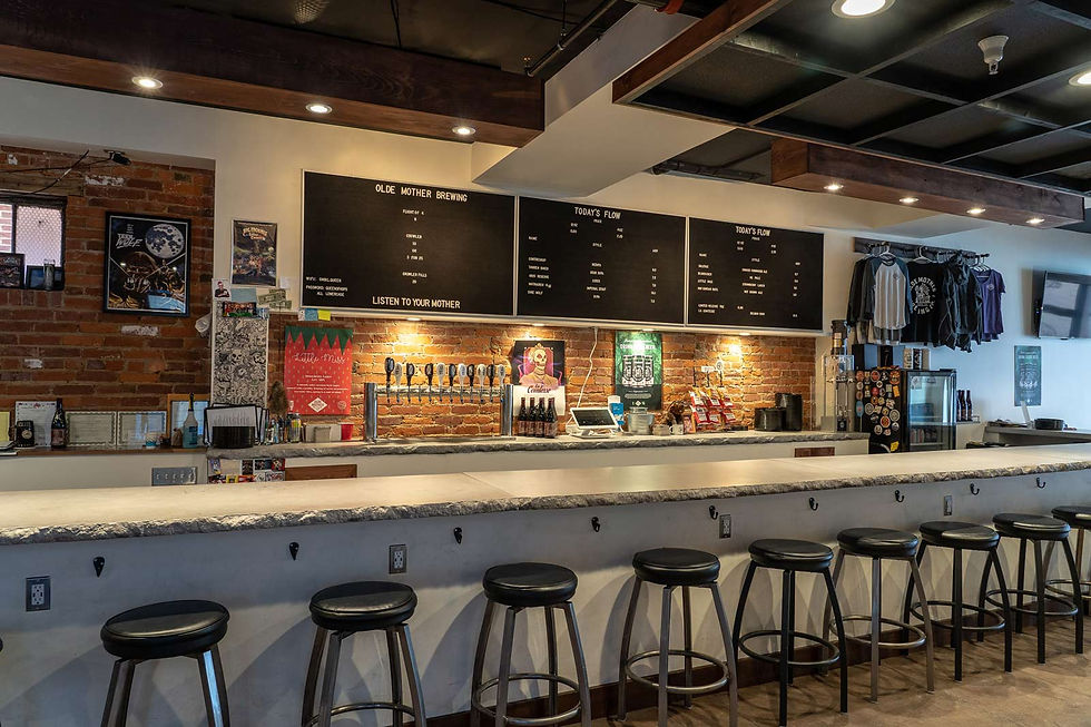 Industrial-style bar with eight stools, brick wall, beer taps, and shirts for sale. Blackboards list craft brews. Warm, inviting ambiance.