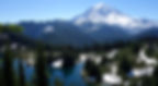 Snow-capped Mt. Rainier with lush pine forest and a clear blue lake. Bright sky enhances the serene, natural landscape.