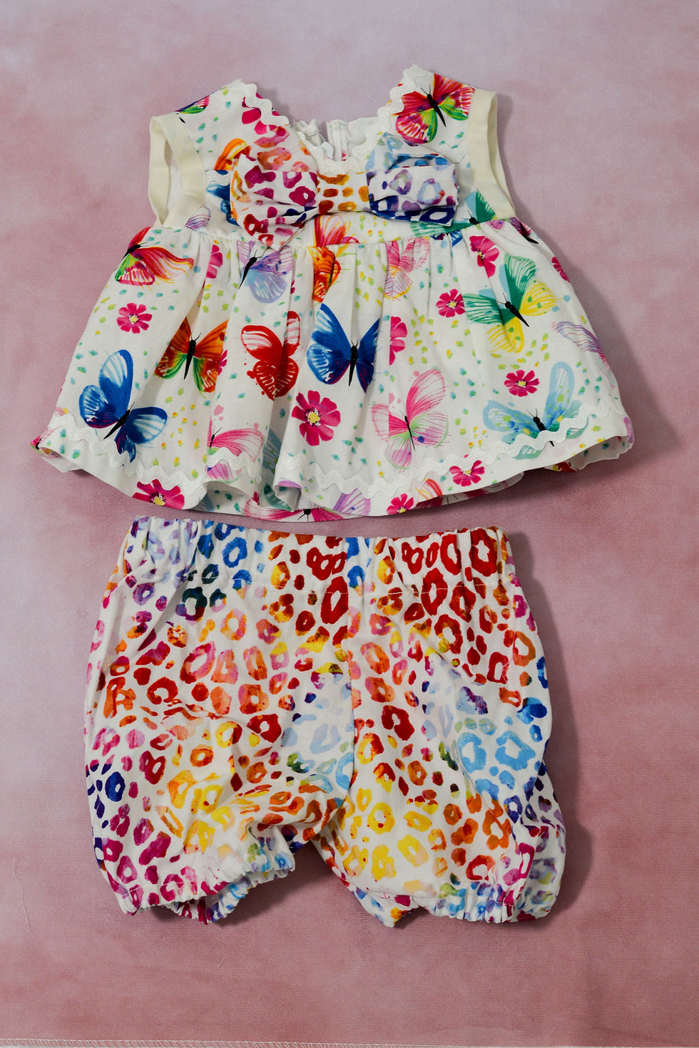Thumbnail: Flat lay of dress/bloomers. Delicate floral pattern on white background. The trim contains white lace and a rainbow pattern.