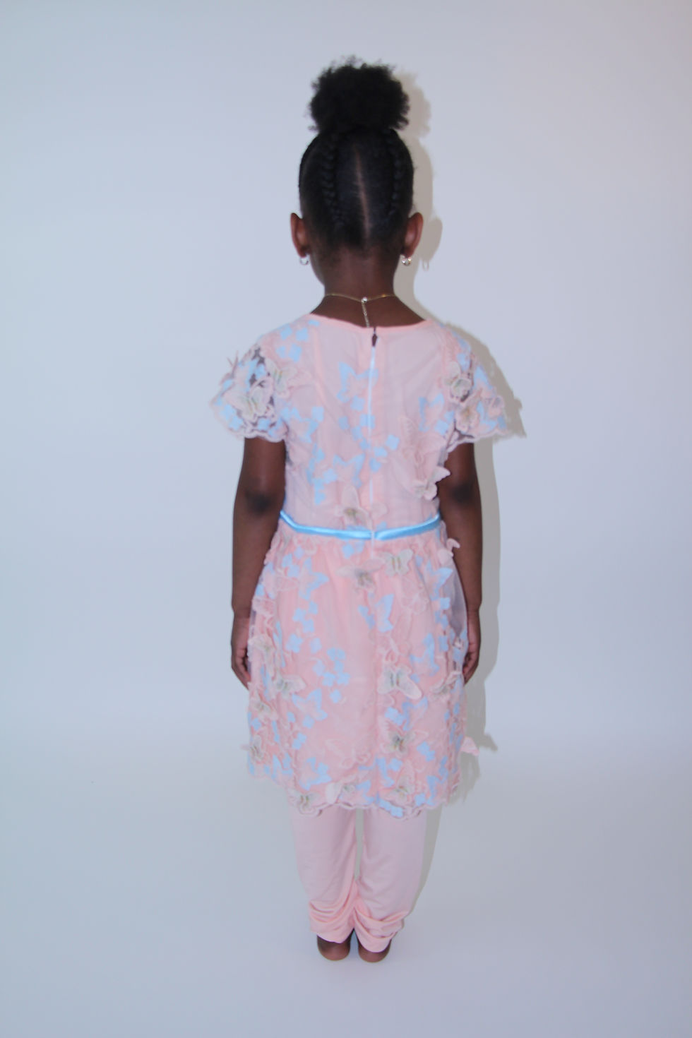 Thumbnail: A young girl in an Olivia Inspirations peach dress adorned and scalloped with blue butterflies and peach leggings back view.