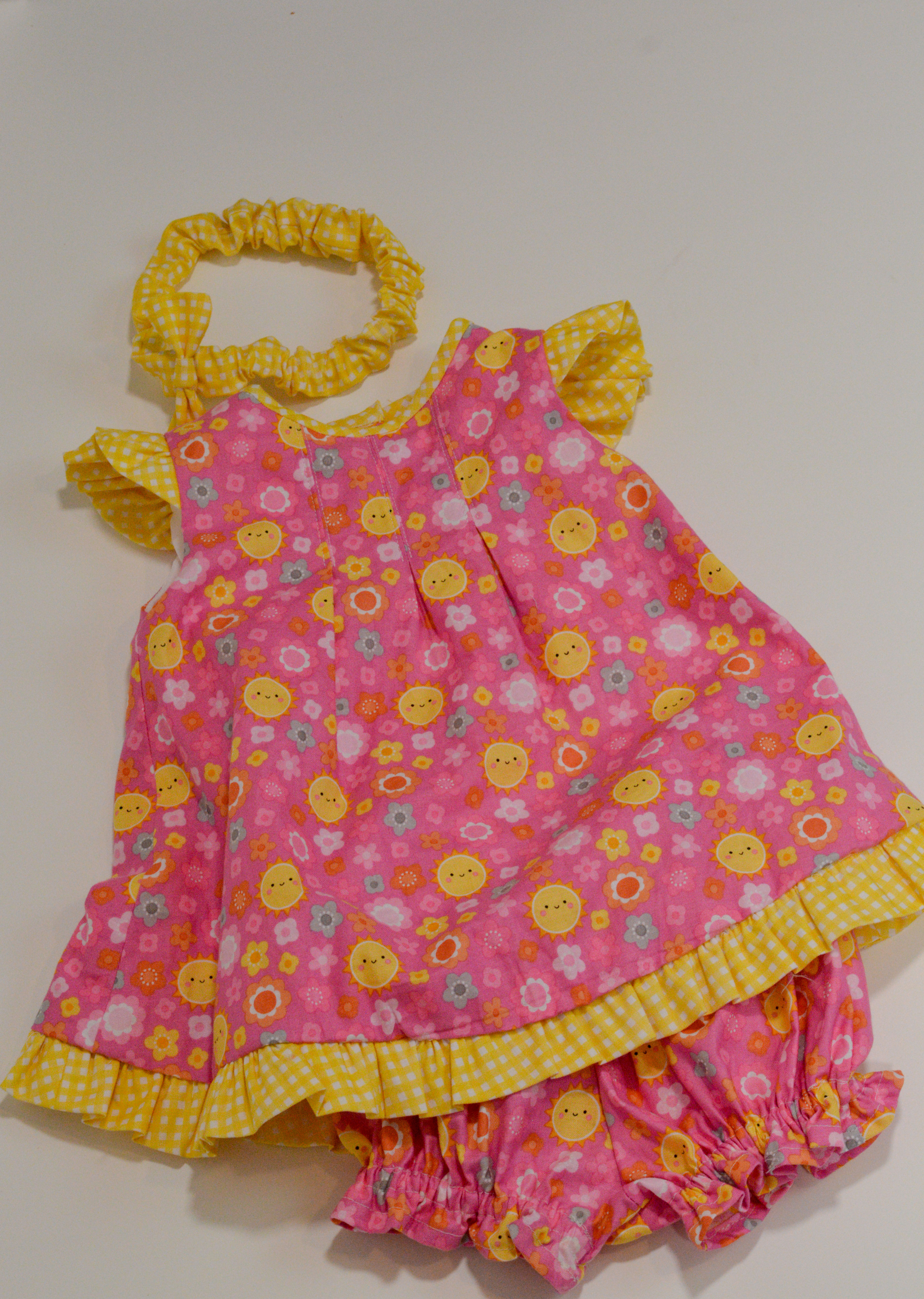 Front view of dress. Pink background with yellow sunshine smiley faces. Trim is yellow gingham.