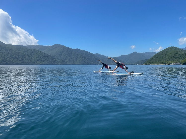 青い湖上で2人がSUPヨガのポーズを取り、背後に緑の山並みと澄んだ青空が広がる、爽快な景色。