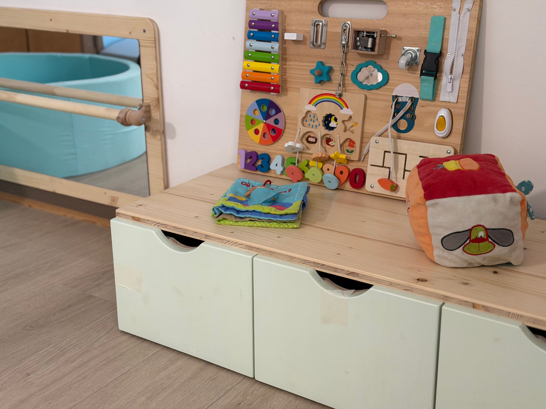 Un panneau d'activités sensorielles en bois posé sur un meuble de rangement bas à la crèche Zarafa, à côté d'un livre en tissu, d'un gros dé d'éveil et d'un miroir de motricité.