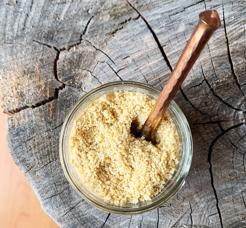 Glass jar of yellow vegan parmesan powder with a bronze spoon on rustic wooden stump. The wood's cracked texture contrasts with the smooth jar.