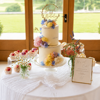 Two-tier semi-naked buttercream wedding cake decorated with pastel fresh flowers and displayed on a styled dessert table with cupcakes at Kingscote Barn wedding venue.