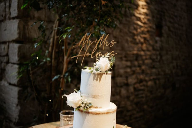 Two-tier semi-naked wedding cake with white roses on a log slice at Cripps Barn, topped with a "Mr & Mrs Holmes" sign.