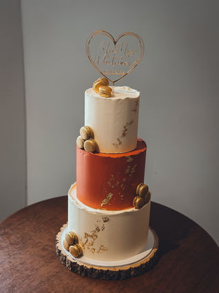 Large three-tier buttercream wedding cake with burnt orange centre tier, gold macarons and leaf detailing, on a rustic wood slice at The Kings Head, Cirencester.