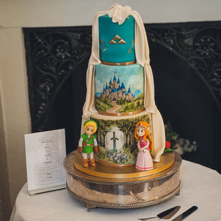 Three-tier hand painted Zelda wedding cake featuring Hyrule Castle, the Master Sword, Triforce symbol, fondant draping, gold details and handmade Link and Princess Zelda figures.