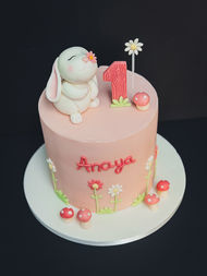 Blush pink tall first birthday cake with handmade fondant bunny topper, red number one, daisies, toadstools and the name Anaya on the front.