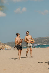A group of people are gathered in a bright, wooden-floored yoga studio, sitting on yoga mats in a relaxed and cheerful atmosphere. An instructor sits at the front, smiling and engaging with the group. The room has stacks of neatly arranged blankets, cushions, and yoga props against the back wall, with natural light streaming in through open windows.