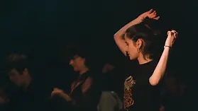 Woman with dark hair tied up, wearing a black T-shirt with a dragon graphic, raising her arms while dancing or stretching, with other blurred people in the background on a dark stage or studio.