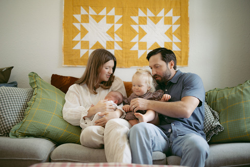 Family of four on a couch, parents holding a newborn and toddler, smiling in a cozy living room with a yellow quilted wall hanging. Considering the best time for newborn photos in your home