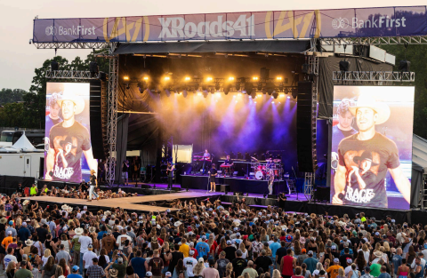 Crowd watches outdoor concert stage lit purple and gold, with large screens showing a performer and XRoads4, BankFirst banner.