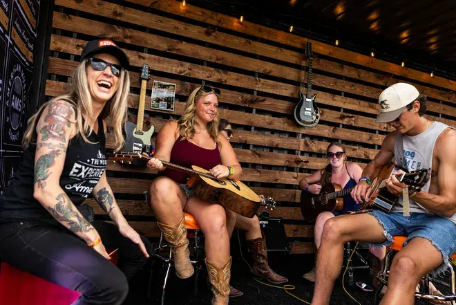 Smiling musicians jam with guitars in a rustic bar with wood slat walls and hanging guitars.