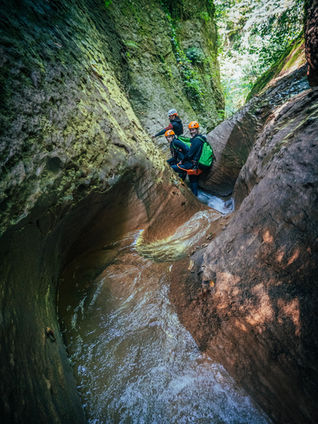 A small group of adventurers wearing helmets, wetsuits, and green backpacks navigate a narrow, flowing river section inside a deep, rocky gorge at Dzmuisi Canyon.