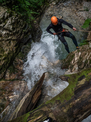A high-angle view of a canyoneer wearing a black wetsuit and orange helmet carefully rappelling down a rocky cliff next to a rushing, white-water waterfall in Dzmuisi Canyon.