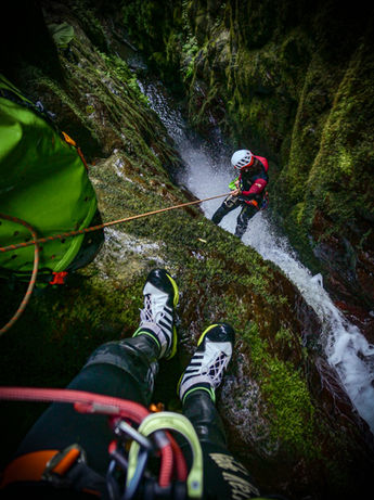 A first-person view of a canyoneer sitting on a steep, mossy cliff edge with safety ropes and canyoning shoes visible, looking down at another adventurer rappelling down a waterfall into a deep forest gorge at Sopho Canyon.