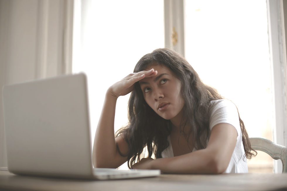 Woman in white shirt leaning on desk, hand on forehead, looking frustrated. Laptop in front, bright window behind, calm setting.