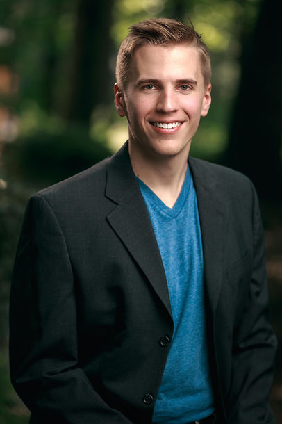A headshot of a male team member dressed in a black suit and a blue shirt. The team member has a confident and approachable expression. The headshot is set against a nature background, showcasing a harmonious blend of professional attire and the beauty of the natural environment.
