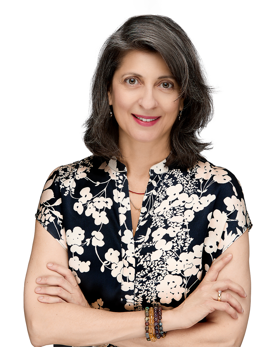 Woman with medium-length gray hair and a floral-patterned blouse smiles with arms crossed. Wears beaded bracelets. White background.