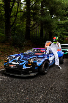 Driver seated on a colourful race car surrounded by woodland.