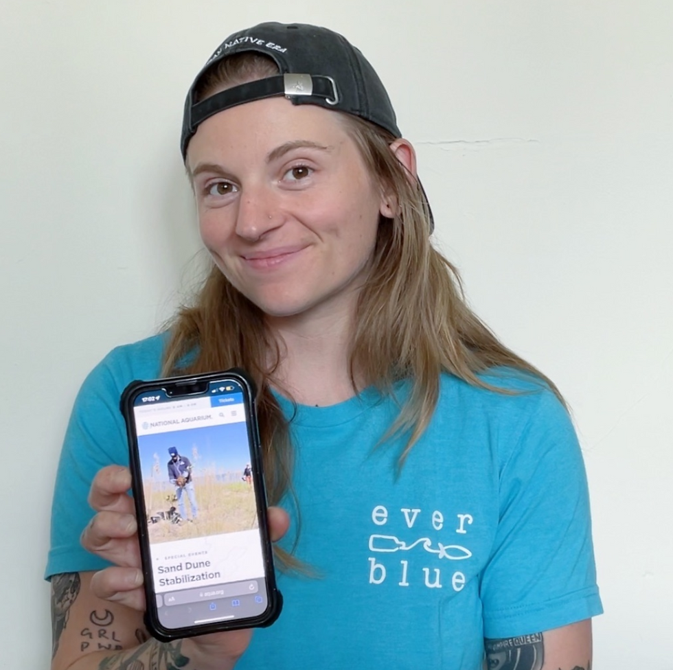 Team member Sibly holds up their phone to a website showing sand dune stabilization volunteer event that they are attended at the end of May 2023, hosted by The National Aquarium.