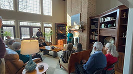 Wide shot of a community gathering in a bright living room with tall windows and a brick fireplace. A man stands near the fireplace speaking to a seated group of attendees arranged around the room on couches and chairs. Sunlight fills the space, highlighting wooden bookshelves, plants, and home décor.