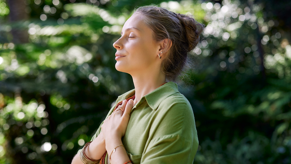 Woman in a green shirt, eyes closed, hands on heart, in a serene forest setting with sunlit foliage. Calm and peaceful mood.