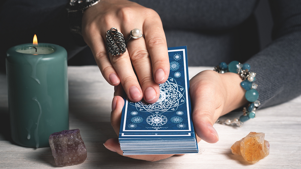 Hands holding a deck of tarot or oracle cards over a table, surrounded by crystals and a lit candle, suggesting a spiritual or divination practice.