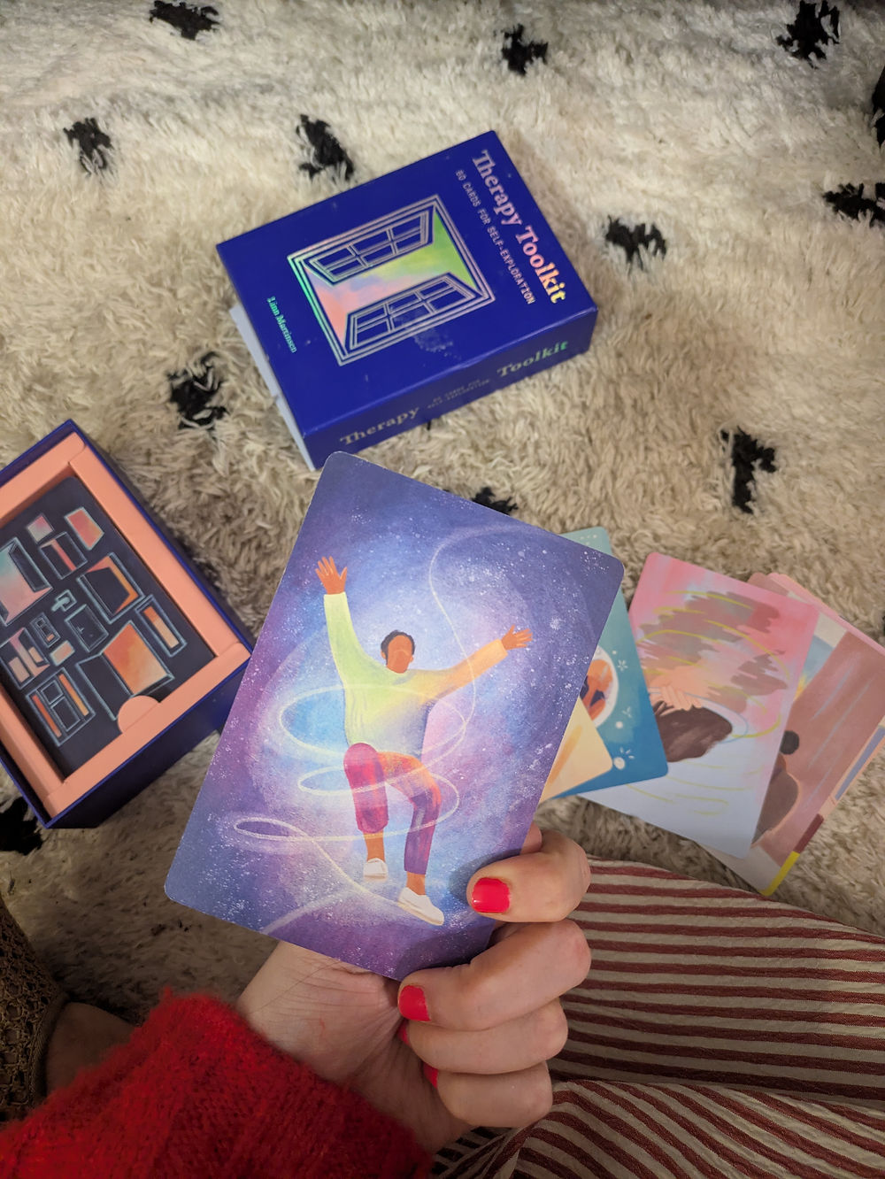 an array of illustrated cards laid out on the floor and Roxanne holding one in her hand. The cards have different types of pictures on, all illustrated with colours and drawings