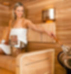 Woman in a towel inside a sauna placing water on the stones to produce steam