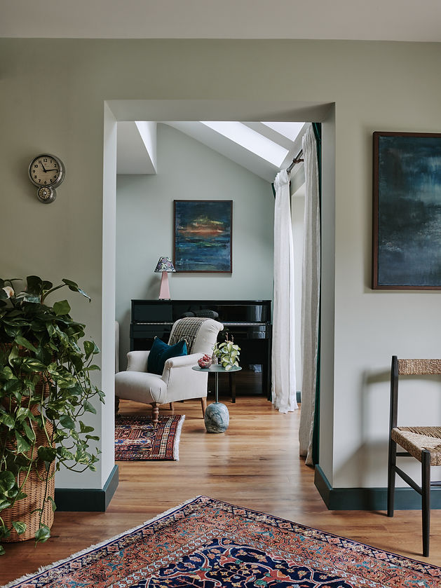 Elegant living space designed by Studio November, featuring a soft armchair with a velvet cushion, a grand piano, and layered Persian-style rugs. The room is styled with natural greenery, abstract artwork, and warm wood flooring in a Cotswolds home.