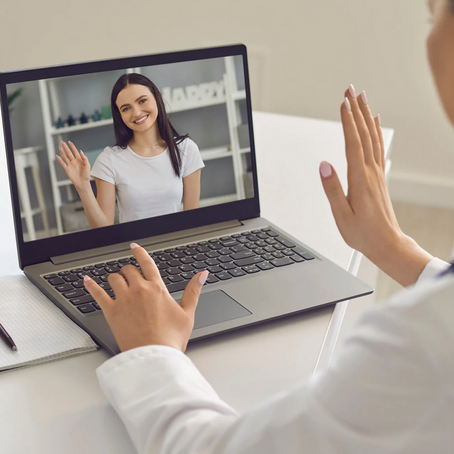 A woman in a white coat sits at a desk, waving toward a person on a laptop screen during a video call.