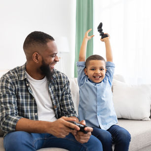 A dad and his young son sitting on a sofa playing video games together. The son is cheering with his arms raised while the dad laughs and holds a controller.