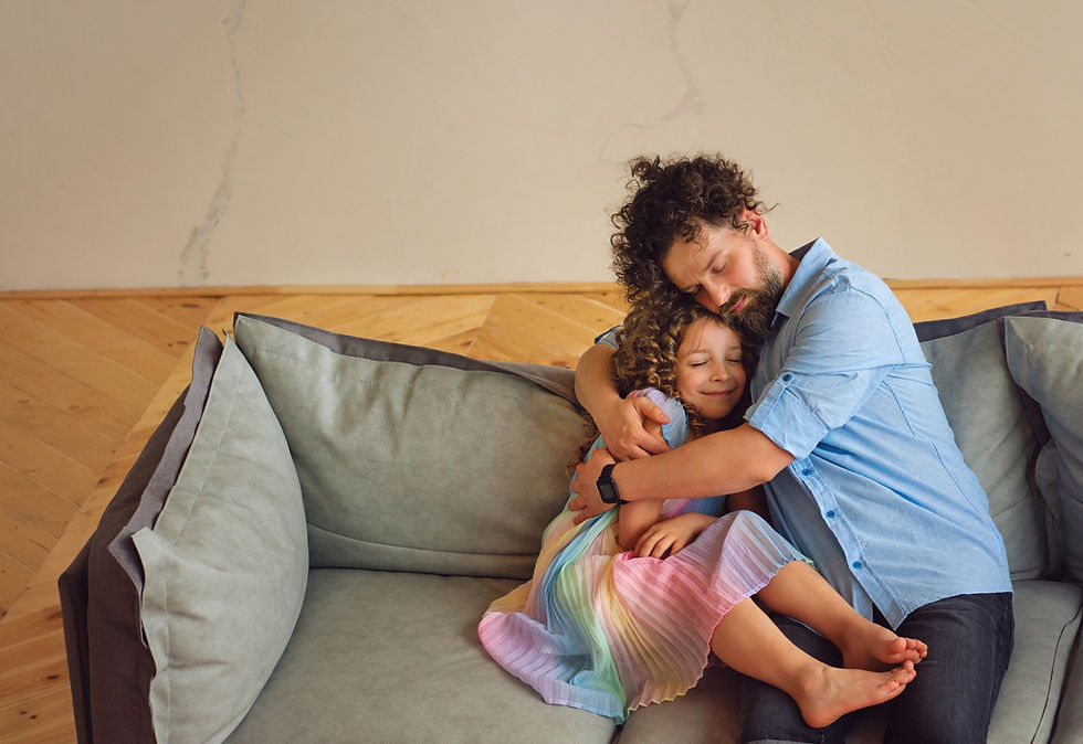 A tired dad and his young daughter cuddle on the couch, both with their eyes closed and relaxed expressions, showing quiet connection and comfort.