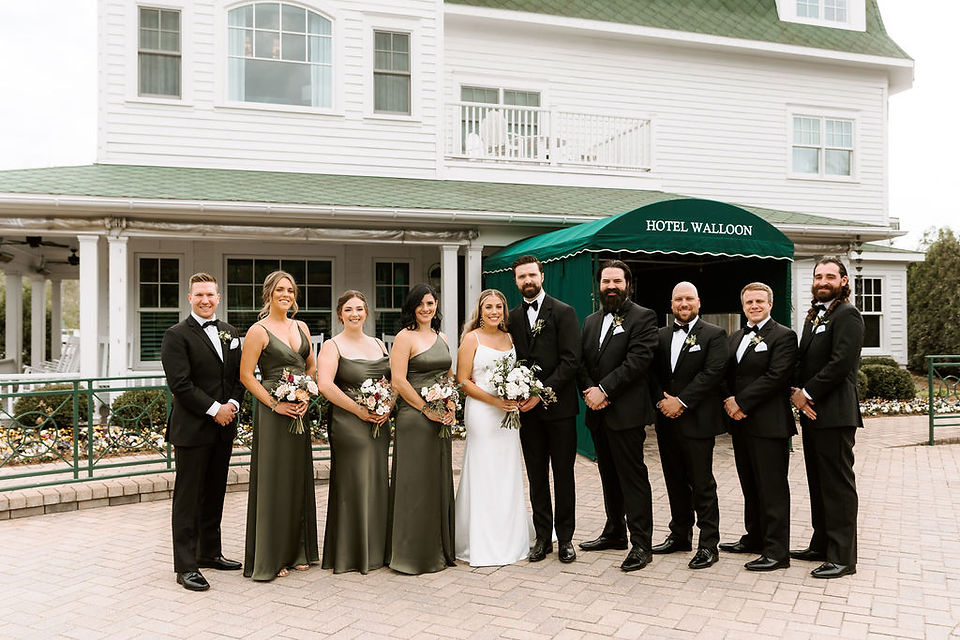 Bridal party portrait featuring sage green bridesmaid dresses and black groomsmen attire in front of Hotel Walloon in MI.