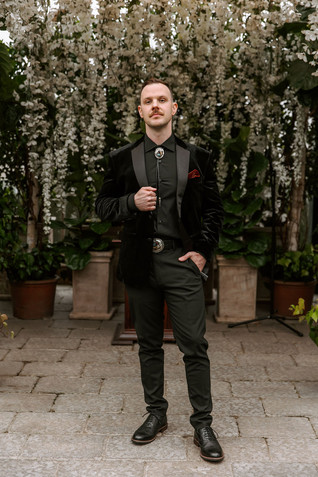Groom in all black attire and bolo tie standing in front of a lush greenery background.