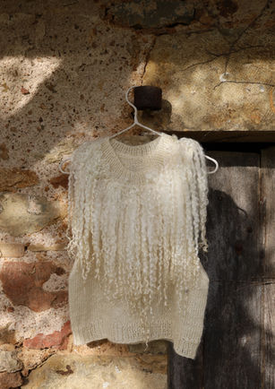 A cream-coloured knitted top by ELWIN. The knitted top is hanging on a wooden beam outside of a farm house against a rugged stone wall, at Chilton Grounds Farm. Image by Mila Burcikova.
