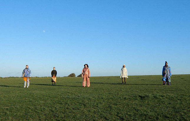 A landscape image of five people standing spaced out on a grassy field with a bright, clea