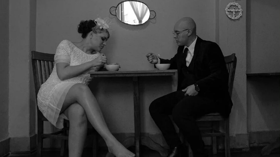 Casal em preto e branco senta-se à mesa, comendo sopa em clima íntimo e sério, numa sala simples com espelho na parede.