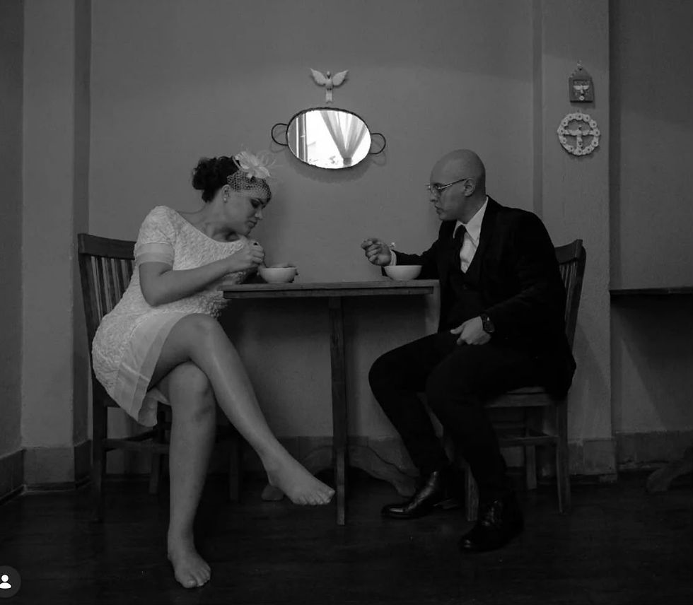 Casal em preto e branco senta-se à mesa, comendo sopa em clima íntimo e sério, numa sala simples com espelho na parede.