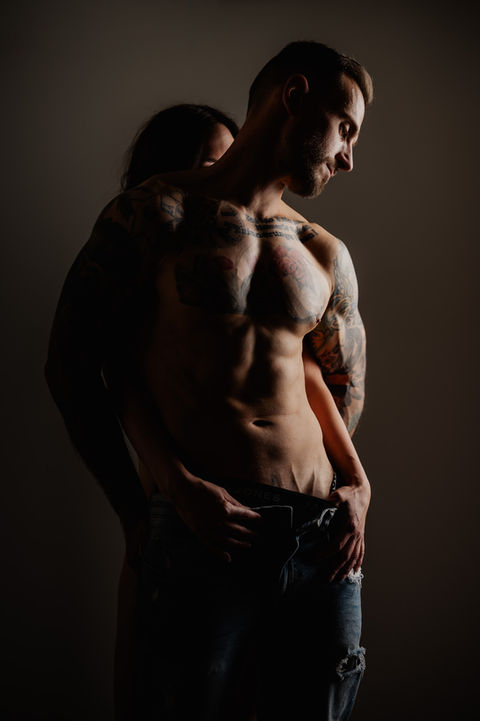 Mann und Frau in Unterwäsche umarmen sich eng, Mann hat tätowierte Arme, stehen in einem dunklen Raum.