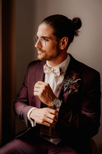 Bräutigam in einem dunkelroten Anzug hält eine Blume und schaut nachdenklich zur Seite.