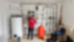 Installateur arbeitet an Heizungsanlage mit Rohren, Pufferspeicher und Steuerungseinheit in Technikraum.