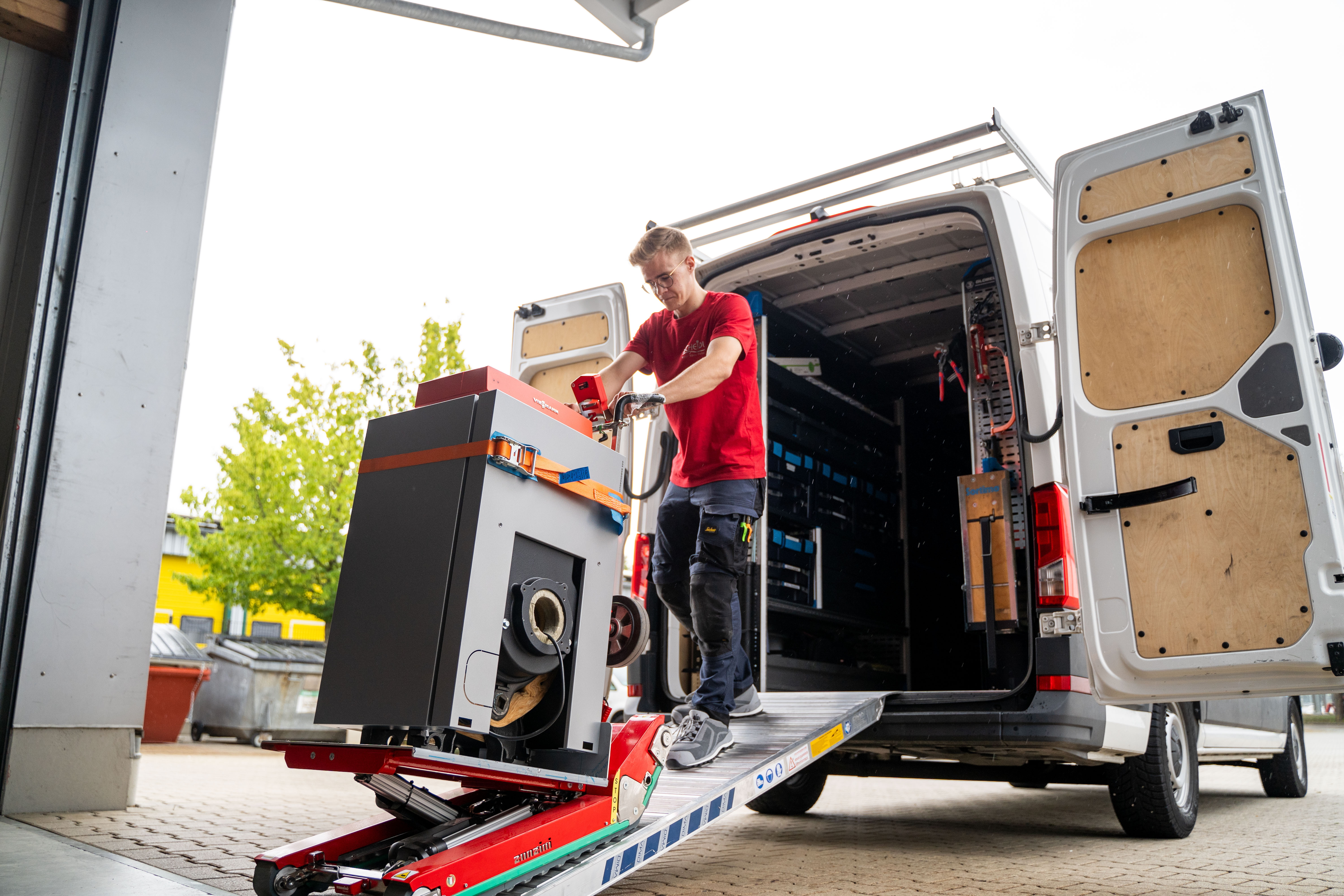 Ein Monteur mit rotem T-Shirt und Brille fährt rückwärts einen Heizkessel mithilfe einer T