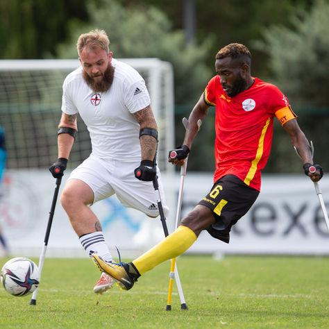 Two amputee soccer players on crutches battle for the ball on a grassy field, one in white and one in red and yellow.